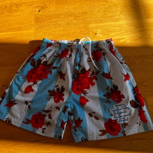 Blommiga shorts från EE - Snygga shorts med blommigt mönster i rött och blått från EE. De har en elastisk midja med snörning för justerbar passform. Perfekta för en avslappnad stil.