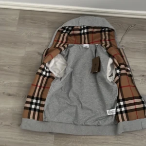 Grå huvtröja med rutig insida - Snygg grå huvtröja från Burberry med klassiskt rutigt mönster på insidan. Tröjan har en dragkedja framtill och justerbar huva med snörning. Perfekt för en stilren och bekväm look. Säljs billigare pågrund av att den inte riktigt passade mig finns även påse ett snöre har blivit lite slitet från posten 