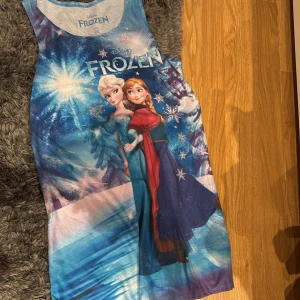 Blå ärmlös klänning med Frozen-motiv - Säljer en blå ärmlös klänning från Disney med motiv av karaktärerna från Frozen. Klänningen har ett vinterlandskap på baksidan och är tillverkad i polyester och spandex. Perfekt för alla Frozen-fans! ❄️( köpte den i USA )