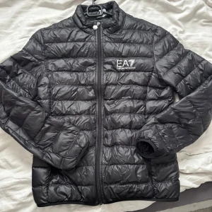 Svart pufferjacka från EA7 Emporio Armani - Säljer en stilren svart pufferjacka från EA7 Emporio Armani. Jackan har en quiltad design med dragkedja framtill och en hög krage. Perfekt för våren. Ej använd!