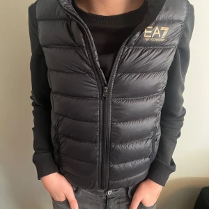 Svart dunväst från EA7 Emporio Armani - Snygg svart dunväst från Armani. Perfekt till våren/sommaren. Finns inget hål eller andra fel på västen, skick 9/10. Säljer västen pågrund av att den är för liten, Storlek 150. Tveka inte på att ställa frågor!
