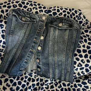 Jeanskorsett från Zara - Snygg jeanskorsett från Zara med knappar framtill och en elastisk baksida för bättre passform. Perfekt för att ge en cool touch till din outfit. Den har en klassisk blå jeansfärg och fransiga kanter för en trendig look.