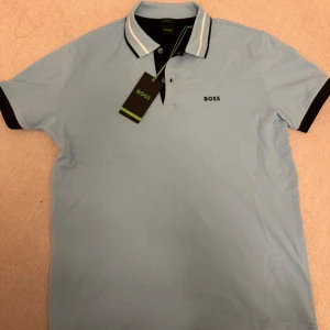 Ljusblå pikétröja från Boss - Snygg ljusblå pikétröja från Boss med svart krage och ärmslut. Tröjan har en klassisk passform med knappar framtill och en liten Boss-logga på bröstet. Perfekt för en stilren look. Helt ny fick den i present men använder ej så den är aldrig använd. Ny modell också och är regulare fit. Helt ny kostar 1100kr men vill bli av med den så säljer billigare 