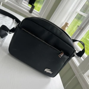 Svart axelväska från Lacoste - Säljer en stilren svart axelväska från Lacoste med silverfärgad dragkedja och klassisk krokodil-logga framtill. Väskan har justerbar axelrem och flera fack med dragkedja för smidig förvaring. Perfekt för att bära med sig det viktigaste.