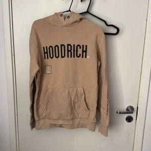 Beige hoodie från Hoodrich - Säljer en beige hoodie från Hoodrich med stor svart logga framtill och tryck på ryggen. Tröjan har huva och en stor magficka. Perfekt för en avslappnad och street-inspirerad stil.