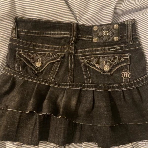 Svart jeanskjol med volang från Miss Me - Säljer en svart jeanskjol från Miss Me med volang och snygga detaljer på bakfickorna. Kjolen har dekorativa knappar och broderade detaljer, samt en kort passform som ger en cool look. Intressekoll kom med bud