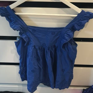 Blå blus med volang från MNG - Säljer en blå blus från MNG med breda axelband och volangdetaljer. Blusen har broderade hålmönster och dekorativa sömmar framtill. Perfekt för varma dagar och ger en söt och somrig look. Går att ha på båda håll, 