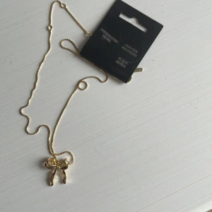 Guldfärgat halsband med rosett från Pilgrim - Snyggt halsband i guldfärg från Pilgrim med ett hänge i form av en rosett. Kedjan är tunn och elegant, perfekt för att ge en söt detalj till din outfit. Halsbandet är tillverkat av minst 75% återvunnet material.orginalpris är 400kr säljer då jag fick det i present efter jag bytigt till silver