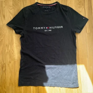 Mörkblå t-shirt från Tommy Hilfiger - Svart t-shirt från Tommy Hilfiger med logga och texten 'EST. 1985' broderad på bröstet. Tillverkad i ekologisk bomull och har klassisk passform med korta ärmar. Diskret flagga på ärmen och ribbad halsringning. Size M