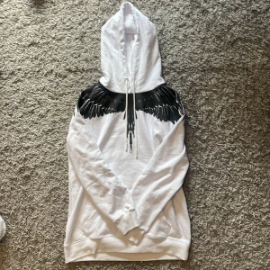 Marcelo burlon hoodie - Säljer en vit hoodie från Marcelo Burlon med ett svart vingmönster över axlarna och bröstet. Tröjan har huva med dragsko och långa ärmar. Perfekt för dig som vill sticka ut med en unik design.