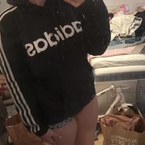 Svart hoodie från Adidas med vita ränder - Säljer en svart hoodie från Adidas med klassiska vita ränder längs ärmarna och stor vit logga på bröstet. Tröjan har huva och magficka, samt långa ärmar. Perfekt för en sportig och avslappnad stil.