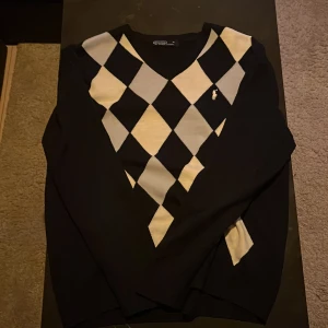 Svart och vit rutig tröja från Ralph Lauren - Snygg svart tröja med klassiskt argylemönster i vitt och grått från Ralph Lauren. Tröjan har v-ringning och en broderad logga på bröstet. Perfekt för dig som gillar preppy stil.