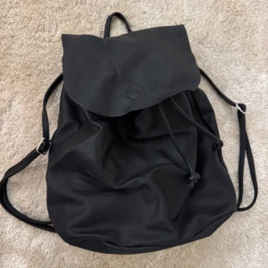 Svart ryggsäck i syntetmaterial - Säljer en stilren svart ryggsäck med justerbara axelremmar och dragskostängning. Väskan har ett lock som stängs över öppningen och är tillverkad i ett slätt syntetmaterial. Perfekt för dig som vill ha en enkel och snygg accessoar till vardagen.