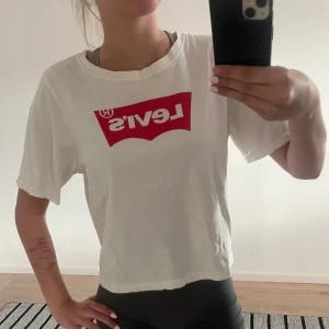 Vit t-shirt från Levi's med röd logga - Vit t-shirt från Levi's med klassisk röd logga framtill. Modellen är croppad och har korta ärmar. Tillverkad i mjuk bomull, perfekt för en avslappnad stil.