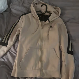 Beige hoodie med dragkedja från Adidas - Snygg beige hoodie från Adidas med dragkedja och huva. Klassiska svarta ränder längs ärmarna och svart panel över axlarna. Perfekt för en sportig look och har två stora fickor framtill.