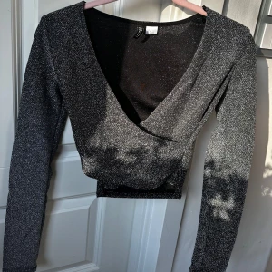 Glittrig omlottopp i svart från H&M - Säljer en svart glittrig omlottopp med lång ärm och croppad passform från H&M. Toppen har djup v-ringning, materialet är stretchigt och skimrar snyggt i ljuset. Använt några gånger ❤️