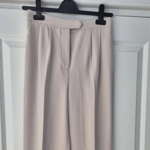 Beige kostymbyxor från NA-KD - Snygga beige kostymbyxor från NA-KD med rak passform och hög midja. Byxorna har vida ben och är tillverkade i ett mjukt, vävt material som ger en stilren look.