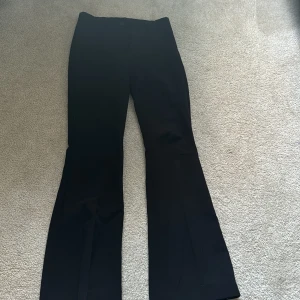 Svarta kostymbyxor med bootcut - Snygga svarta kostymbyxor med bootcut passform och klassiska fickor bak. Byxorna har hällor för bälte och stängs med knapp och dragkedja. Perfekta för en stilren look.