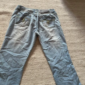 Ljusblå jeans med broderad bakficka - Säljer ett par ljusblå jeans med snyggt gult och vitt broderi på bakfickorna. Byxorna har mer eller mindre bootcut design, lite rakare än vanligt bara. Inga skavanker eller defekter förutom loggan där bak som gått sönder i tvätten, detta syns på bilderna men är verkligen ingenting som stör. De är i storlek L men passar mer som s/m