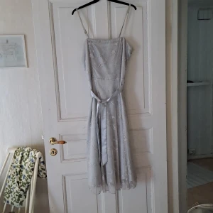 Ljusblå långklänning med broderade detaljer - Säljer en ljusblå långklänning med smala axelband och broderade blommor. Klänningen har ett avtagbart bälte i midjan och är tillverkad i ett lätt, transparent material med foder under. Perfekt för sommarens tillfällen!