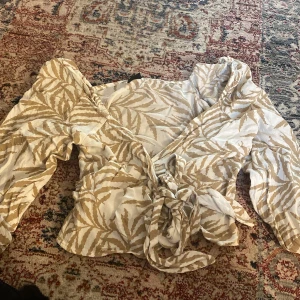 Beige och vit omlottblus med knytband - Säljer en snygg omlottblus i beige och vitt med bladigt mönster. Blusen har långa ärmar med lätt puff och knytband i midjan. Perfekt för dig som gillar trendiga detaljer och mönster.