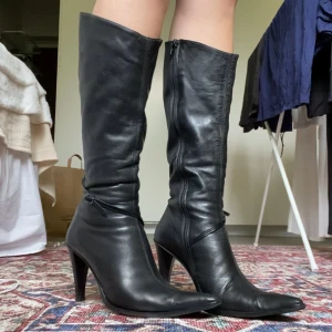 Svarta höga stövlar med klack - Säljer ett par svarta höga stövlar i skinn med spetsig tå och hög klack. Stövlarna har dragkedja på insidan och en smal remdetalj runt vristen. Passar en 39. Står storlek 40 men funkar även för mig som har 38. Väldigt fint och mjukt skinn, bra dragkedja och skönt foder. Klack ca 10cm.