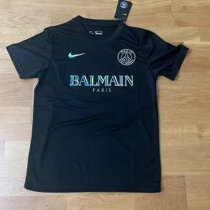 PSG fotbollströja från Nike x Balmain - Snygg svart PSG fotbollströja i samarbete med Balmain. Tröjan har korta ärmar, rund hals och holografiska detaljer på logga och sponsortryck. Tillverkad i lätt och ventilerande Dri-FIT material för bästa komfort på planen.