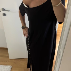 Svart klänning med knappar - Svart klänning med asymmetrisk offshoulder-design och dekorativa knappar längs ena sidan. Klänningen har en lös passform och är tillverkad i ett mjukt material som faller snyggt. Perfekt för dig som gillar en stilren och modern look.