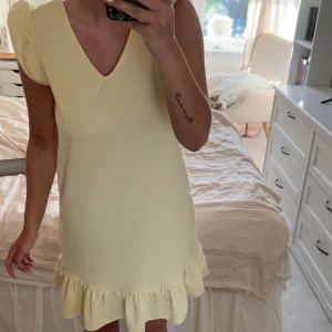 Ljusgul klänning med volangdetaljer - Söt ljusgul klänning med volang på axlarna och nederkanten. Klänningen har v-ringning fram och en djupare v-ringning i ryggen. Perfekt för varma dagar och har en feminin, lekfull stil.