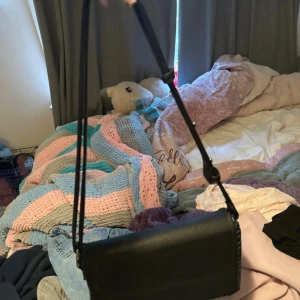 Svart axelväska i skinnimitation - Säljer en stilren svart axelväska med lång rem och lock. Väskan har enkel design och är tillverkad i skinnimitation. Perfekt storlek för det viktigaste och passar till många olika outfits.