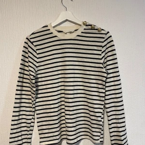 Randig långärmad tröja med guldknappar - Säljer en vit och svart randig långärmad tröja med tre dekorativa guldknappar på ena axeln. Tröjan har rund halsringning och är i ett mjukt, stretchigt material. Perfekt för en stilren och klassisk look.