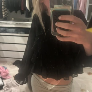 Svart glittrig blus med volanger från Gina Tricot - Säljer en svart blus från Gina Tricot med glittriga detaljer och flera lager volanger framtill. Blusen har långa, transparenta ärmar och är croppad i modellen. Perfekt för dig som vill sticka ut lite extra!