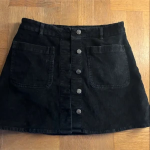 Svart manchesterkjol med knappar - Svart kjol i manchestertyg med raka linjer och två stora fickor framtill. Kjolen har knappar längs hela framsidan och hällor för bälte. Perfekt för dig som gillar en enkel men snygg stil.