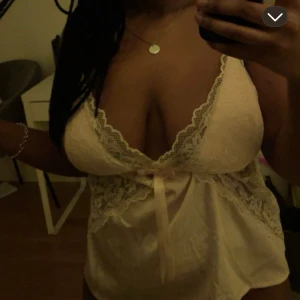 Ljusrosa spetslinne med rosett - Säljer ett ljusrosa linne med spetsdetaljer och söt rosett framtill. Djup v-ringning och tunna axelband, perfekt för en feminin look. Mjukt material och genombrutna spetspartier ger en romantisk känsla.