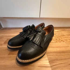 Svarta Loafers - Svarta vintage unisex loafers i storlek 40. Skorna har dekorativa fransar och brogue-detaljer framtill, samt en robust sula med kontrastsöm. I mycket bra skick, knappt använda. Perfekta för en stilren vardagslook eller dressat avslappnad stil. Säljer då de var för små för mig. Nyskick