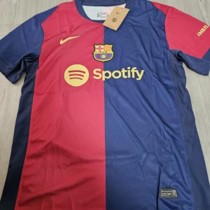 FC Barcelona hemmatröja 2023/24 - Säljer en FC Barcelona hemmatröja från säsongen 2023/24 med Spotify som sponsor. Tröjan är röd och blå, har klubbmärke och Nike-logga på bröstet samt UNHCR-tryck på ryggen. Klassisk design med korta ärmar och rund hals.