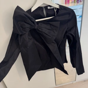 Svart topp med stor rosett från NA-KD - Säljer en svart långärmad topp från NA-KD med en stor dekorativ rosett på axeln. Toppen har rund halsringning och är tillverkad i ett glansigt material som ger en elegant look. Perfekt för att sticka ut i mängden!