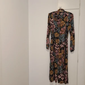 Blommig långklänning från Monki - Säljer en långklänning från Monki med färgglatt blommönster i rosa, blått och gult på svart botten. Klänningen har lång ärm, krage och knappar framtill. Perfekt för dig som gillar retrovibbar och färg.