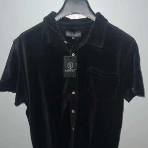 Svart kortärmad skjorta från LAGENCY Svart/beige Finns  - Snygg svart skjorta från LAGENCY med kort ärm och klassisk krage. Skjortan har knappar framtill och en bröstficka. Perfekt för en stilren look. Svart skjorta i storlek m och beige skjorta I L