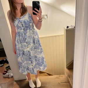 Vit och blå klänning med knappar - Säljer en söt vit klänning från H&M med blått mönster och knappar framtill. Klänningen har smala axelband, v-ringning och ett romantiskt tryck. Köptes på H&M i Grekland! Perfekt för varma dagar och har en luftig passform som ger en avslappnad känsla.
