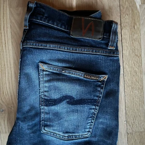 Mörkblå jeans från Nudie Jeans - Säljer ett par mörkblå jeans från Nudie Jeans med klassisk femficksdesign och snygga kontrastsömmar. Jeansen är i slim passform och är tillverkade i slitstarkt denim. Perfekta för dig som gillar stilrena och tidlösa jeans.