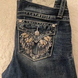 Miss Me jeans med paljettfickor - Säljer ett par blå Miss Me jeans med snygga detaljer på bakfickorna, dekorerade med paljetter och broderi i silver och beige. Jeansen har normal passform och klassisk bootcut-stil. Dom är midwaist och använda kanske max 10 ggr. Köptes från usa för ca 2300 med frakt midjemått: 75, innerbernslängs: 77! ❤️❤️