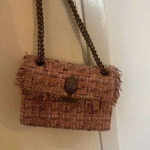 Rosa axelväska från Kurt Geiger - Säljer en snygg rosa axelväska från Kurt Geiger London. Väskan har ett grovt vävt tyg med inslag av guldtråd och fransiga kanter. Kedjerem i metall och dekorativt spänne framtill. 