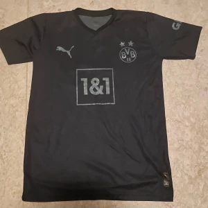 Dortmund Fotbollströja  - Säljer en svart Borussia Dortmund fotbollströja från Puma med klubbmärke och sponsortryck på bröstet. Tröjan har korta ärmar och V-ringning. Perfekt för dig som vill visa ditt stöd för BVB.