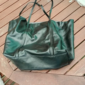 Svart shoppingväska i syntet - Stor svart shoppingväska från H&M i syntetmaterial med dubbla axelband. Väskan har en enkel design och ett rymligt huvudfack med innerficka. Perfekt för att bära med sig allt man behöver till skolan eller stan.