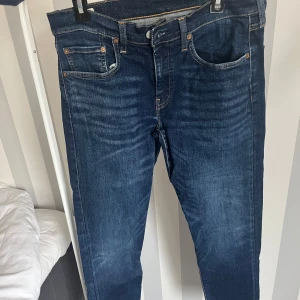 Levi's 502 mörkblå jeans - Säljer ett par mörkblå Levi's 502 jeans med klassisk femficksdesign och orange sömmar. Jeansen har normal passform och raka ben, tillverkade i slitstarkt denim. Perfekta för dig som gillar tidlös stil och vill ha ett par jeans som funkar till det mesta.