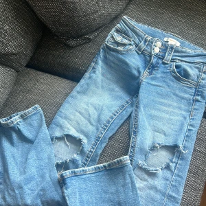 Blå  jeans med hål ifrån Gina Tricot - Säljer ett par blå jeans från Gina Tricot i storlek 146. Jeansen har slitna hål på båda knäna, klassisk femficksmodell och dubbla knappar i midjan. Perfekta för en avslappnad och trendig look.