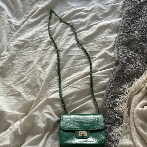 Grön axelväska med krokodilpräglat mönster - Säljer en snygg grön axelväska med krokodilpräglat mönster och guldfärgat spänne. Väskan har en kedjerem med gröna detaljer och ett stilrent, kompakt format. Perfekt för att lyfta din outfit!