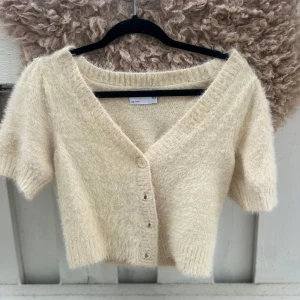 Beige fluffig kofta med v-ringning - Supermjuk och fluffig beige kofta med kort ärm och v-ringning. Koftan är croppad och har knappar framtill. Ribbstickade detaljer vid ärmslut och nederkant. Skriv om ni vill ha bild på (vid matchning av kläder). SKRIV INNAN KÖP!❤️