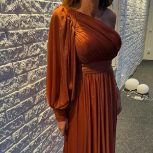 Glittrig rostorange långklänning med oneshoulder - Säljer en elegant rostorange långklänning med glittrigt tyg och oneshoulder-design. Klänningen har en lång puffärm och markerad midja som ger en snygg siluett. Storlek 34/36 hör av dig för fler bilder 